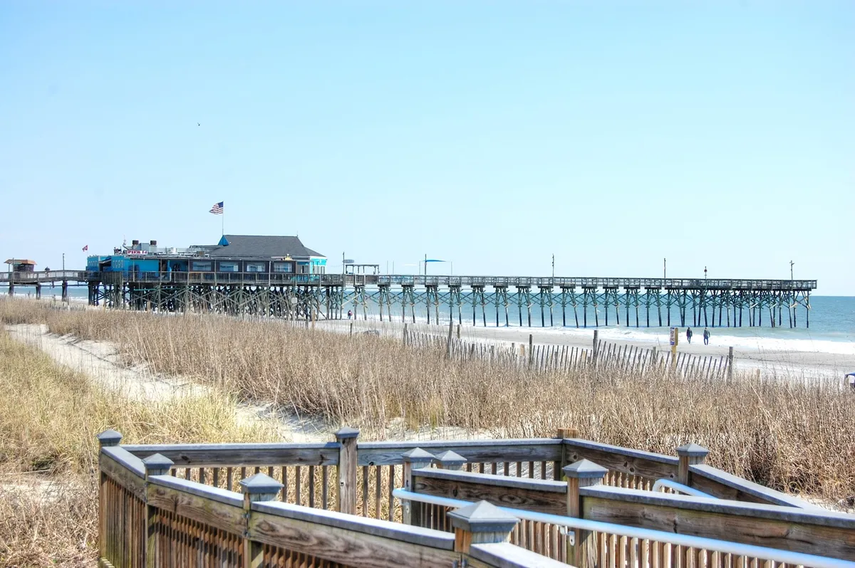 A classic Myrtle Beach pier stretching over the ocean, surrounded by dunes, beach sand, and vibrant blue skies. The scene is tranquil, with gentle waves and an open invitation to stroll along the shore or enjoy the sunset.