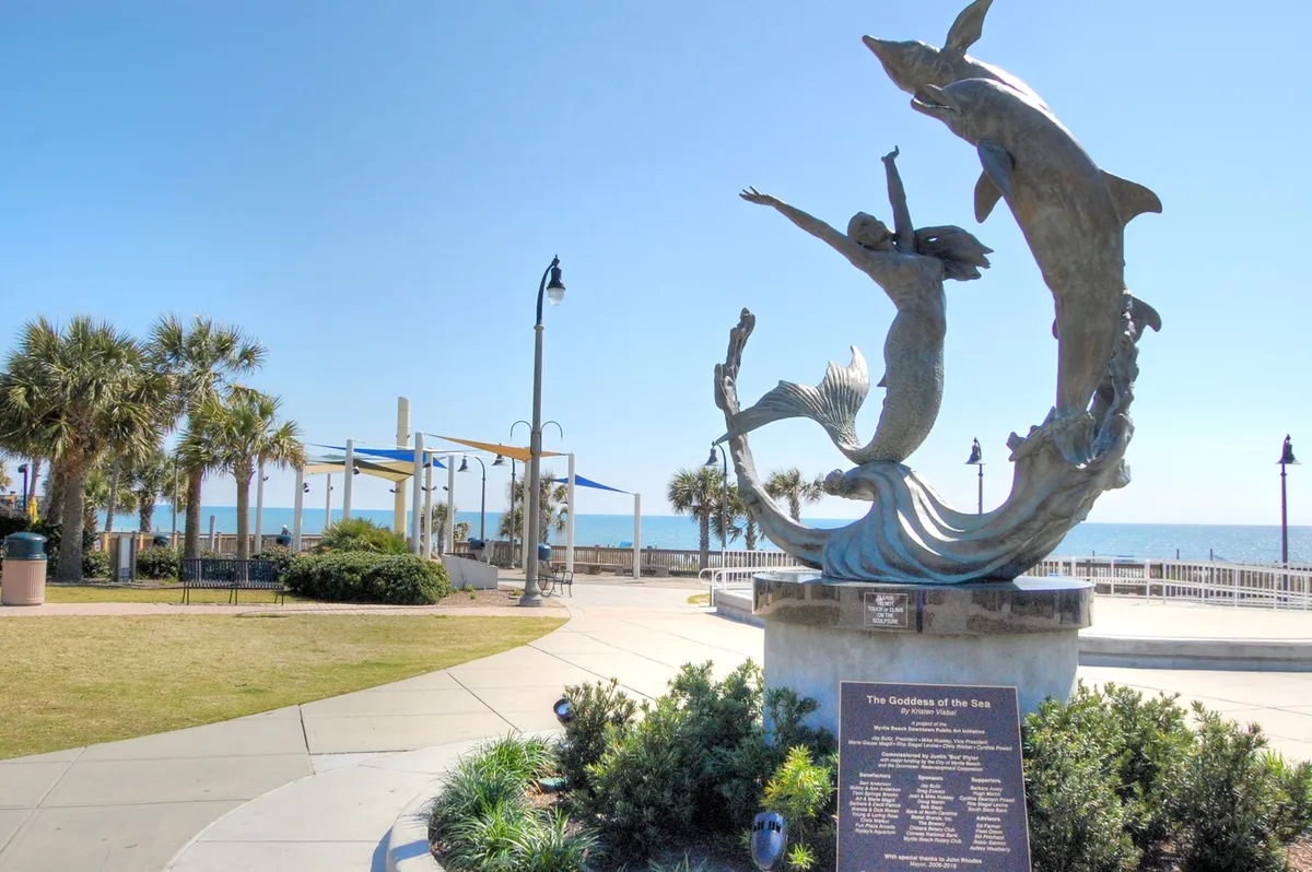 A picturesque park scene with a mermaid and dolphins sculpture set against an ocean backdrop, framed by palm trees and open spaces. The area feels serene yet playful, ideal for leisurely strolls or memorable photo opportunities.