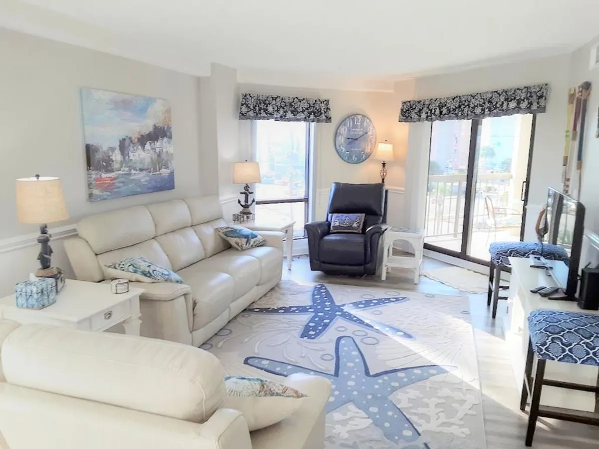This image displays a bright, open living room with a cream-colored sectional sofa, a blue accent chair, a large starfish-patterned rug, and framed coastal artwork. The TV stand with a TV is visible, and sunlight streams through the windows, giving the space a cheerful, beachy ambiance.