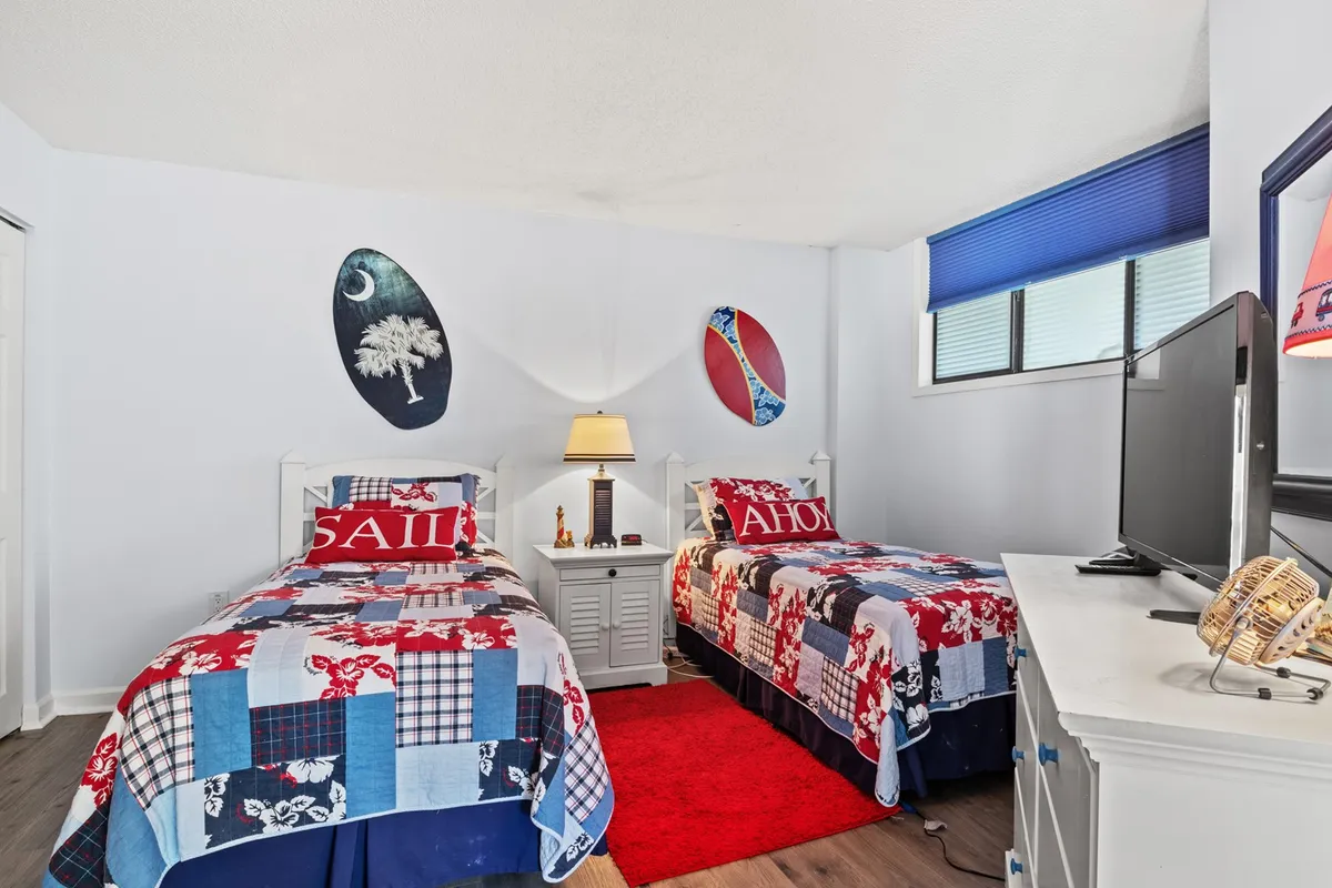 A playful twin bedroom featuring two beds with colorful quilts, surfboard wall decorations, and a cozy nightstand with a lamp. The room exudes a fun, inviting vibe ideal for children, friends, or family members sharing the space.