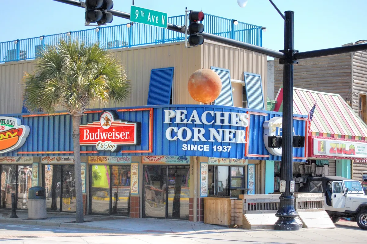 A bright exterior shot of the well-known Peaches Corner with its vibrant signage, Budweiser sign, colorful awnings, and the bustling street scene. Groups of people are visible enjoying the lively local dining scene, which is central to the Myrtle Beach experience.