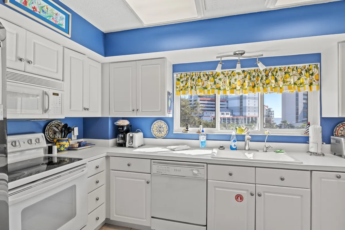 A cozy, uplifting kitchen scene with a blue accent wall, yellow patterned curtain, modern appliances, and a window view. The area feels both functional and inviting, with a focus on everyday ease, whether preparing a meal or enjoying a casual coffee.