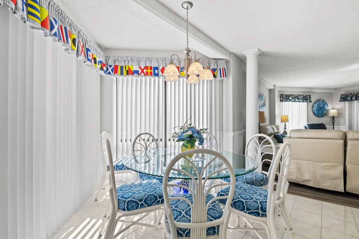 A welcoming dining area with a glass dining table, comfortable chairs, a blue floral centerpiece, stylish chandelier, and a vibrant valance. The dining area is contiguous with a seating area, echoing an open-concept design.
