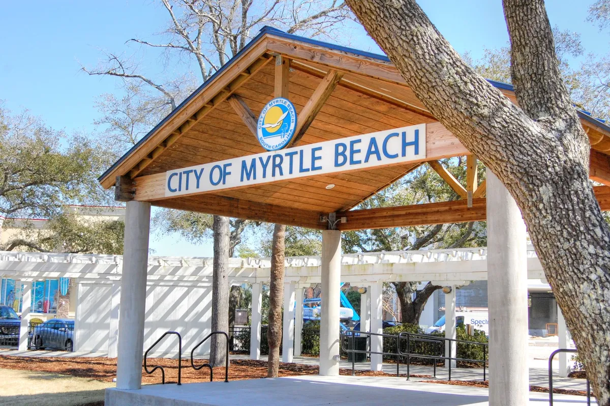 This exterior shot features the welcoming signage of a wooden community structure surrounded by lush trees. The scene conveys a sense of arrival, community engagement, and a peaceful, organized environment.
