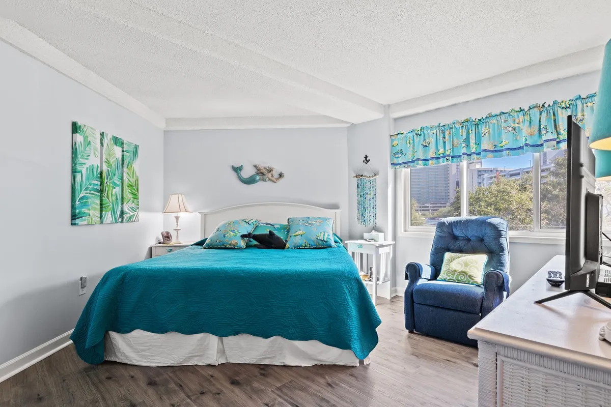 Showcasing a bedroom with teal bedding, tropical-themed wall art, a comfortable blue recliner, and a window with an inviting view. The atmosphere is calm and cheerful, ideal for unwinding, reading, or enjoying entertainment.