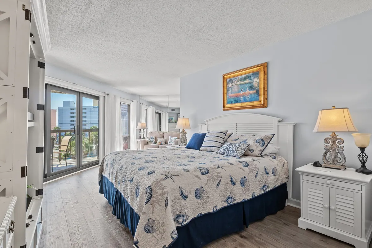This image features a spacious bedroom with a large bed dressed in a blue and white quilt, a cozy lounge area with chairs, and large windows that invite ample natural light and offer scenic views. The room radiates a sense of tranquility and comfort, accentuated by the soft color palette and plush furnishings.