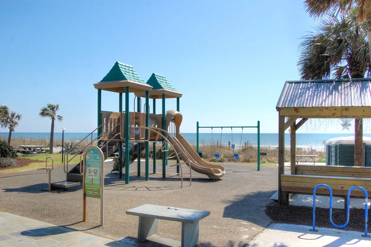 This vibrant exterior image showcases playground equipment and swings with a clear beach view and palm trees in the background. Children can be seen playing while families enjoy picnics, all set against the backdrop of the sunlit shoreline.