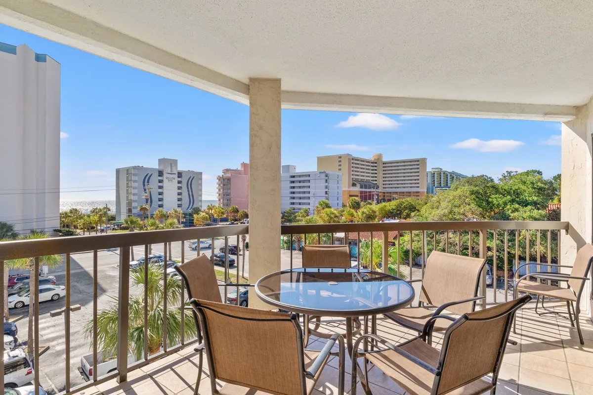 The image features a roomy balcony with comfortable seating, overlooking surrounding buildings and lush palm trees. It conveys an atmosphere of relaxation, with hints of ocean or pool views visible in the background.