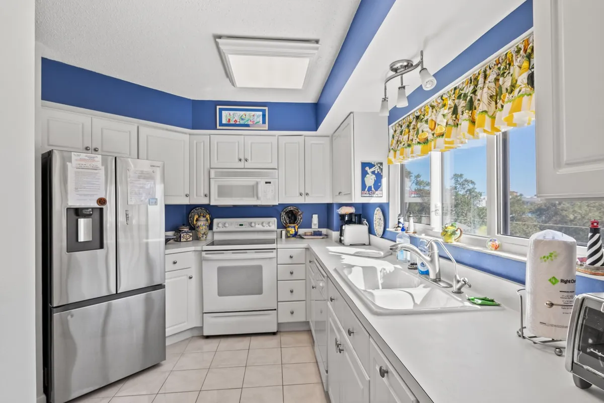 A cheerful and welcoming kitchen featuring white cabinetry, bright blue walls, large windows that let in lots of light, yellow floral curtains, and a stainless steel refrigerator. The scene is lively, emphasizing a space ideal for cooking, baking, and gathering with family or friends. Ample counter space and modern appliances are visible, supporting home-cooked meals and casual entertaining.
