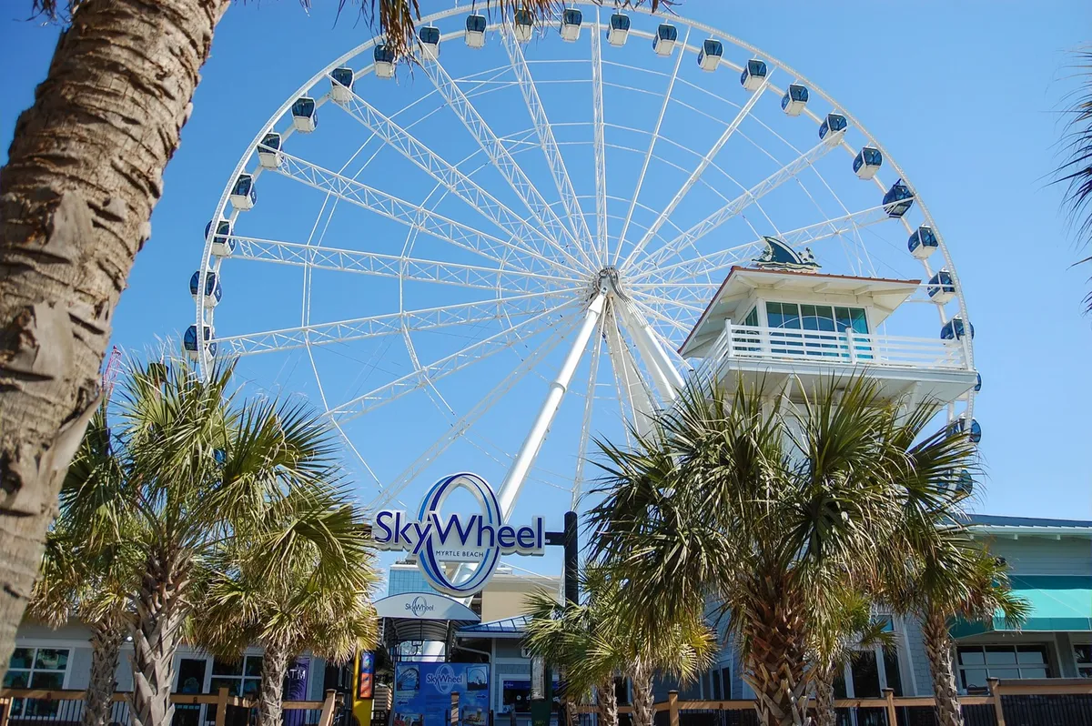 A lively exterior shot featuring the iconic Myrtle Beach SkyWheel, surrounded by palm trees and a ticket booth, with families and groups enjoying the thrill and panoramic views along the bustling boardwalk.