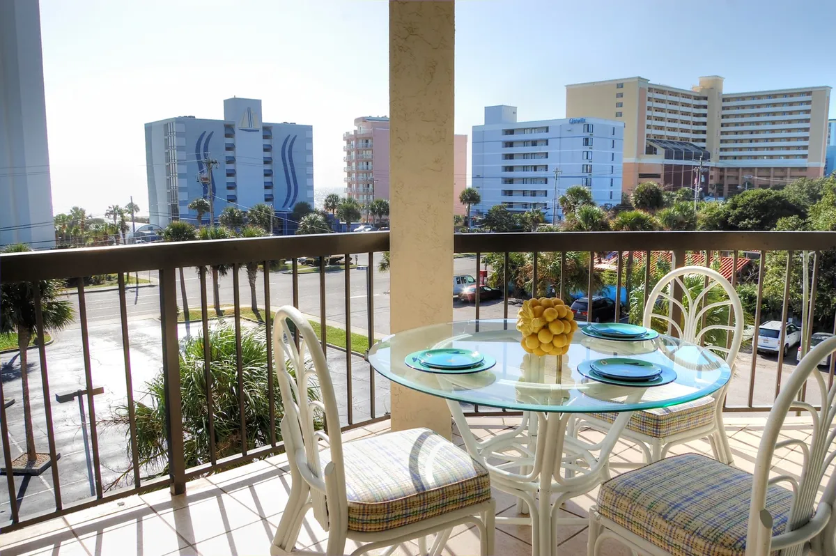 A bright and inviting balcony setup featuring a glass-top table surrounded by colorful chairs, with a cheerful bowl of lemons as the centerpiece. The balcony overlooks the condo's exclusive full-size swimming pool and offers a partial view of the ocean with neighboring buildings in the distance—ideal for outdoor dining, leisurely mornings, and evening relaxation.